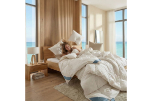 In een Japandi slaapkamer met uitzicht op zee ligt een vrolijke vrouw onder prachtige donzen Bedaffair dekbedden en ze gooit de donzen kussens van Bedaffair omhoog. Ze ligt op het zwevende massief wild eiken Amsterdam bed van Bedaffair.