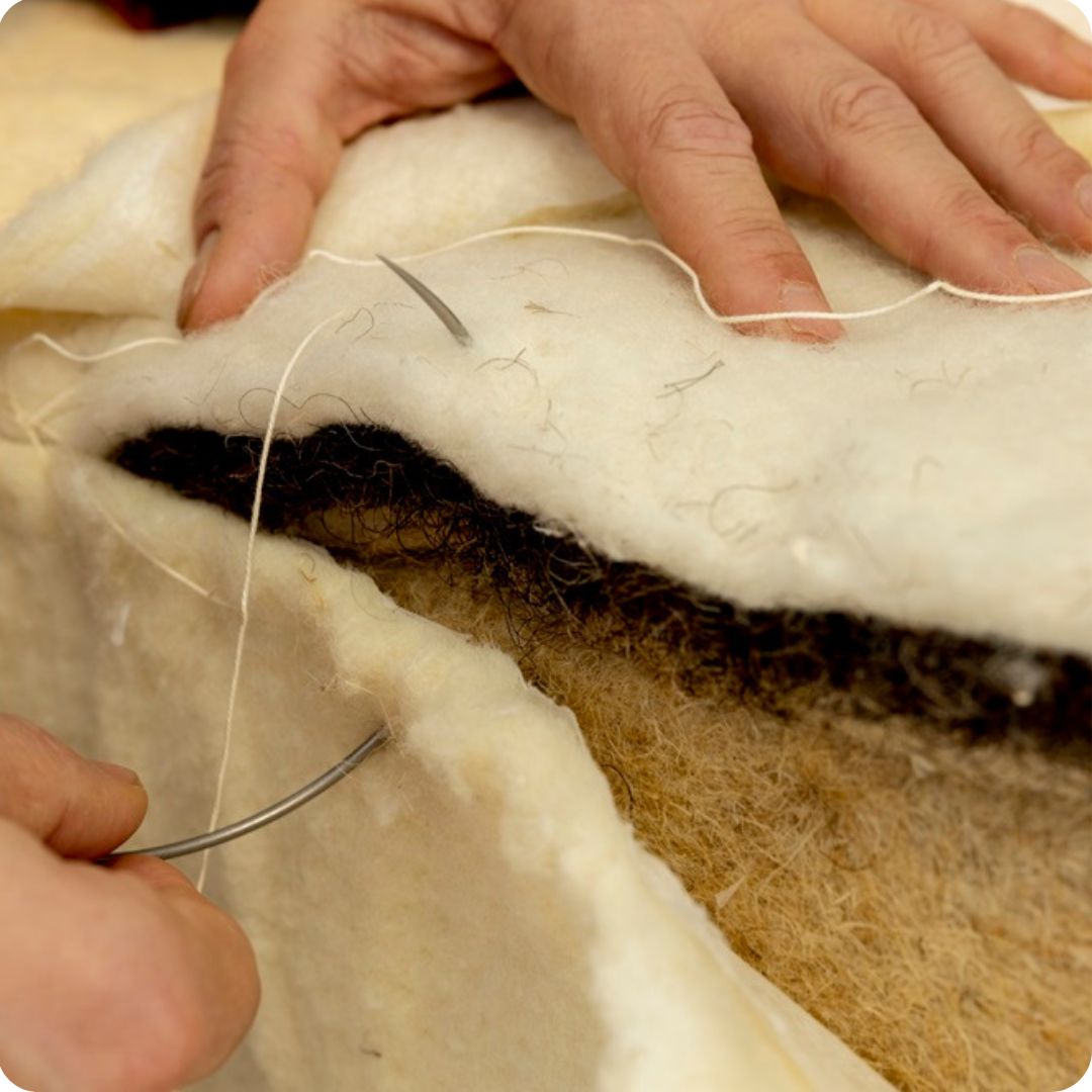 Een close-up foto van de handen van de beddenmaker die met een ronde naald de natuurlijke materialen biologische wol, Moosburger paardenhaar en biologische hennep vezel handmatig dicht aan het naaien is